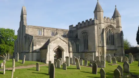 Calre Strak The outside of the church. The sky is blue with no clouds, the grass around the church is green and there are several gravestones on it. The church is a light stone colour and has large glass windows