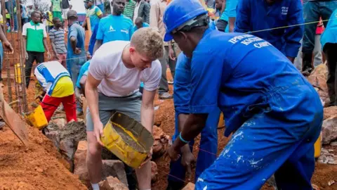 Getty Images Sam Billings unloads some cement into a large hole at a dig, assisted by two Rwandan men in blue construction helmets and uniform. A crowd of men with spades watch or work behind.