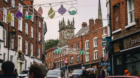 J&PR Wyle Cop in Shrewsbury. There are red brick buildings on either side of a road that has cars driving down it. There are unlit Christmas lights hanging above the street, and people walking around. There is a church in the background. 