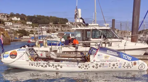 RNLI Penlee A wide shot of a small boat covered in stickers, the words "world's toughest row" are visible at the front