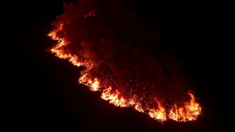 Athena Picture Agency Picture of an orange glowing wall of grass fire ripping across a mountain in the darkness. Flames can be seen tearing through grass, with clouds of smoke rising behind it. 