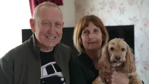 A middle-aged couple stand side by side in front of a TV screen in a living room. The woman is holding a cockapoo. 