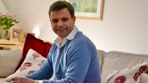 Handout A man in a cornflower blue jumper and white shirt. He is wearing a silver watch and 