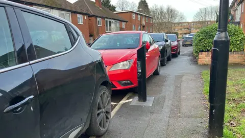The image shows an EV charging point installed on a street in Coventry. Non-electric cars are parked on the road, blocking access to the charger