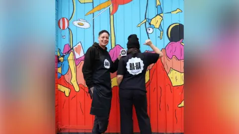 BA-HA Two people in black clothes branded with the word 'BA-HA' stand in front of a colourful mural. One looks at the camera while the other has their back to the camera and raises their arm in the air.