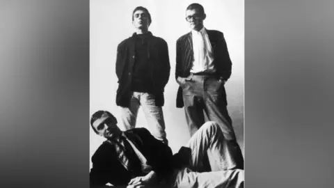 Geoff Reeve/Courtesy of the Phillips family In a black and white image Peter Phillips stands next to artist David Hockney. In front of them Peter Crutch lies on the floor. They are all wearing dark jackets. 