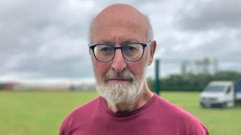 JOANNA TAYLOR/BBC Tony Forster looking at the camera with a glum expression. He is wearing glasses and a dark red T-shirt. Behind him is the partially built green metal fence and a van on a field.