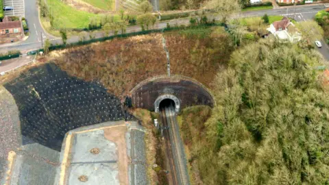 Network Rail A view of the other side of the tunnel in March 2022. The aerial view shows the steep sides of the cutting and buildings dotted around the tunnel. A road goes over the tunnel and there are junctions on both sides.