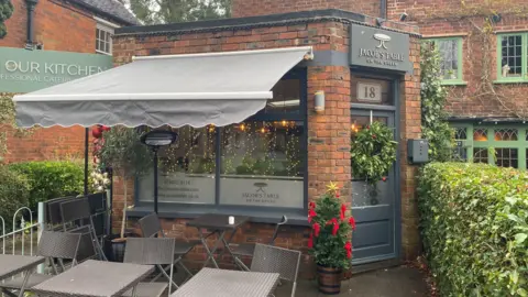 The exterior of Jacob's Table. It is a small, smart-looking cafe with grey colours. There is a green hedge running along the side and some chairs outside. 
