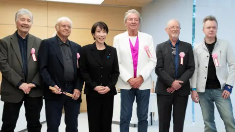 Reuters Takaichi poses with Deep Purple. They stand side-by-side in a line and smile. Left to right are: Don Airey, Ian Paice, Sanae Takaichi, Ian Gillan, Roger Glover and Simon McBride. They are stood in a room with wood-panelled walls, and grey blinds drawn over a windowed wall to the right of the frame.