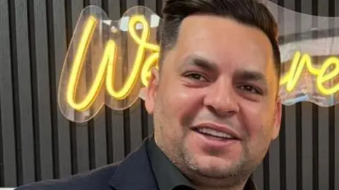 Camran Butt Camran Butt, who has dark brown hair and is wearing a dark blue suit jacket over a black shirt with an open collar smiles into the camera. 