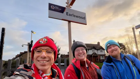 Alex Manners Three young men at Earlswood Railway Station. Stood in front of the station sign on the platform is a man with yellow and purple glasses and red hat and scarf, a man with red coat, glasses and black hat and a man with a blue jacket on, ginger beard and turquoise beanie hat 