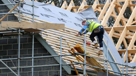 PA Media A roofer on a roof of a house with planks of wood and frame of a roof.