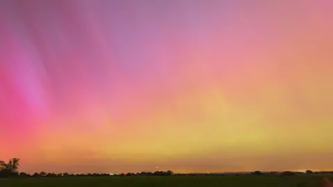 Rik Freeman The northern lights pictured from a field. The sky is purple and pink at the top of the image and yellow and orange towards the bottom. The outline of a field, with houses in the distance fills the bottom of the image. Stars can also be seen in the sky. 