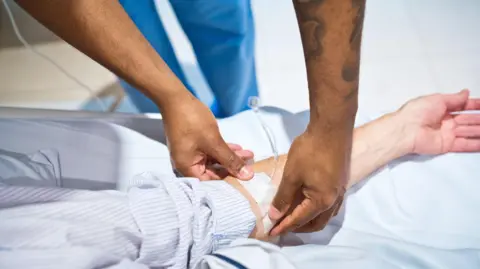 Getty Images A healthcare worker securing medical tape over an IV tube on a patient's forearm. The patients is wearing a long-sleeved striped shirt and their arm rests on a hospital bed.