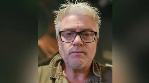 A man with white hair, dark glasses and a green jacket is looking at the camera.