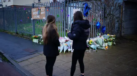 PA Media Two girls standing in front of a vigil for Max and Mason, where flowers are laid against a railing and a stop knife crime banner