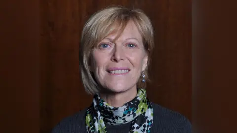 Hilary Knight A woman with sandy coloured hair smiling into the camera. She is wearing a blue jumper and a brightly coloured scarf, and the background looks like wood panelling