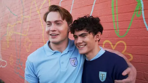 Ann Gannon / BBC Actors Kit Connor and Joe Locke smile smile at the camera, they're both in their Truham High PE kits from the show. Kit has his arm around Joe.