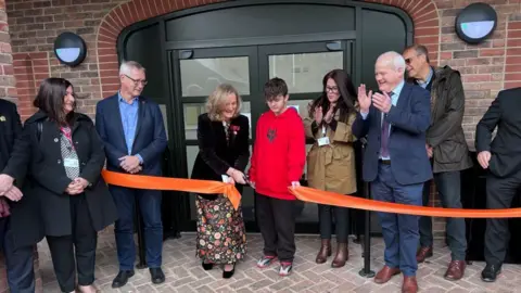 Kate Bradbrook/BBC Nine people, standing in front of a building, that is made out of red brick, by an orange ribbon, that has been cut. They are standing by double doors, one man is clapping and one woman in the middle is leaning over and holding some scissors. 