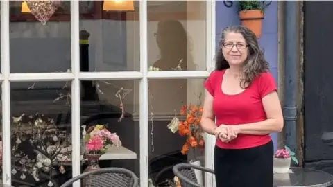 Jane Talbot Jane Talbot wearing a red top outside her Cafe 50. She has long dark hair and is wearing glasses