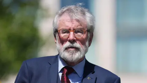 Gerry Adams, with grey hair and a grey beard, wearing a navy suit, blue shirt and red tie. He is wearing round orange glasses. The background of the image is blurred. 