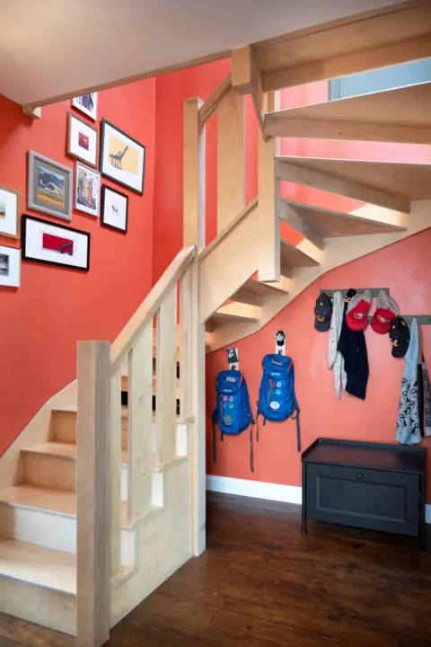 IWC Media A wooden staircase with open risers climbs against bold orange walls, creating a striking, modern interior focal point.
Framed artwork decorates the wall, while hooks below hold jackets and backpacks above a small storage bench on polished wooden flooring.