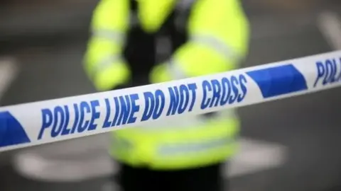 Blurred out shot of a police officer standing behind a cordon. There is blue and white police tape in the foreground.