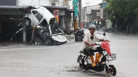 AFP via Getty Images A man wearing a bike helmet, short-sleeved shirt and orange wellington boots rides his scooter past a damaged car that is upturned and leaning on the side of a building in Hualien county, Taiwan.