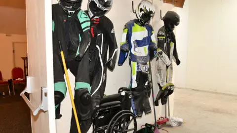 Humberside Police Four sets of motorcycle racing leathers and crash helmets hang from a wall at a funeral parlour. 