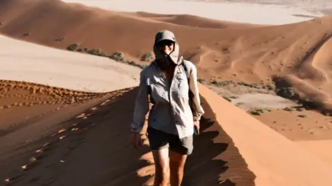 Ellie Mitchell-Heggs Mitchell-Heggs walking in the desert with sand everywhere. He is wearing a sandy coloured hat with side panels and khaki shorts with white trainers. She is smiling at the camera as she walks towards it.