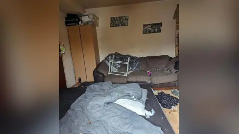 A messy house showing mess and rubbish with a dirty duvet on the floor near a brown sofa.