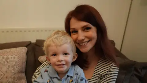 BBC/Peter Whittlesea A woman with dark red hair wearing a striped black and white shirt. She is sitting on a couch and holding her son, who is wearing light blue pyjamas and sitting on her lap. 