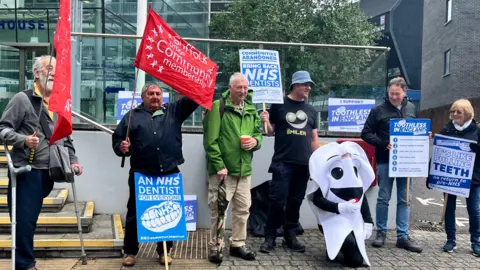 Six member of the Toothless in Suffolk campaign group stand outside Endeavour House. They are equipped with posters and flags.