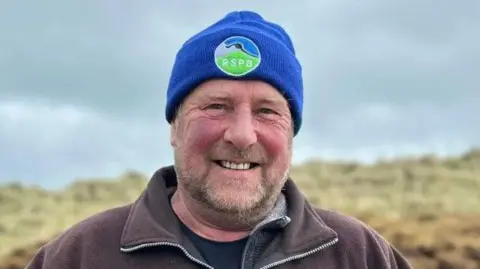 A man standing in a field. He has red rosy cheeks and dark facial hair. He is wearing a blue hat with a 'RSPB' badge. He is wearing a brown zip-up jacket, a grey jacket and a black t-shirt.