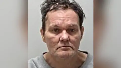 Police mugshot of Adam Saunders, who has brown unruly hair. He is photographed wearing a grey top.