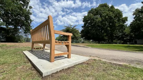 Stuart Woodward/BBC A wooden bench in Chalkwell Park. It is a light brown colour and sits on concrete paving slabs next to a footpath. The bench faces trees and grass
