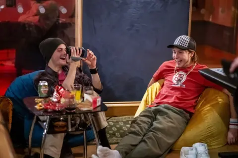 Studio Canal Two young men sitting on colourful bean bags in front of a coffee table littered with snacks. One of the men is using a video camera - both are laughing. They are wearing baggy t-shirts and hats.