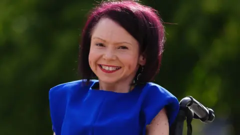 Pam Duncan Glancy beams broadly at the camera out in the garden area of the Scottish Parliament. Her burgundy hair shines in the sun and she wears a bright royal blue dress and a gold bracelet on each arm