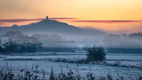 Weather Watchers/Mike Jefferies A view of Glastonbury Tor in the distance as the sun comes up behind the hill. The ground is white and frozen.