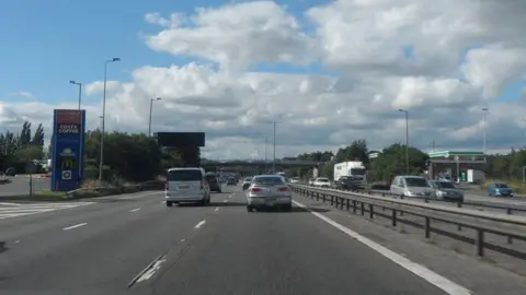 A view of the M1 motorway at Watford Gap Services, as seen from the view of the vehicle. There are a number of cars ahead, on a dual carriageway. 