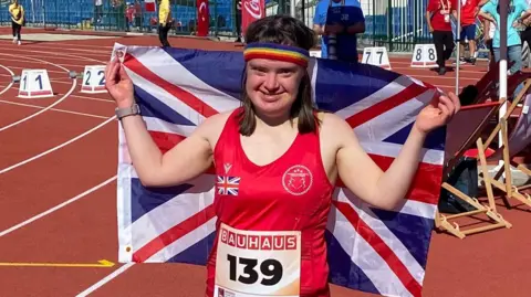 A woman wearing a red sports vest holds a union jack flag in the air. She is standing on a racing track.