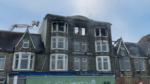 Damaged derelict hotel after a fire. The top floor windows have been destroyed by the blaze. Smoke is coming from the top of the building. The hotel is in a row of other properties. A firefighter can be seen in a cherry picker at the top of the building.