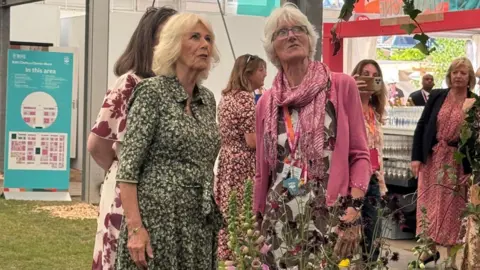 Farewell Flowers Directory Queen Camilla in a button up green and white floral dress standing next to Gill Hodgson, who has a flowery dress on, a pink cardigan and scarf. Gill has short blonde hair and Queen Camilla has shoulder length blonde hair. They are looking up to the right.