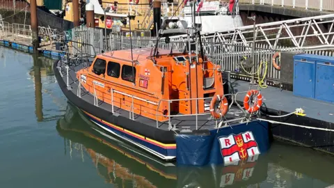 Lowestoft Lifeboat An orange lifeboat is moored next to a dock. It has an RNLI flag on the rear of the vessel. 