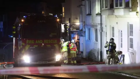 Fire cordon tape across road with fire engine facing up a hill - two firefighters are by the fire engine and two on the path by a white building it is dark. 