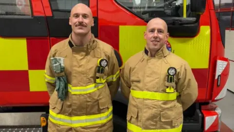 Kieran Thomas Kieran and Andy stand for a photo in front of a red fire truck. They're both bald and wear their biege-coloured firefighter kit.