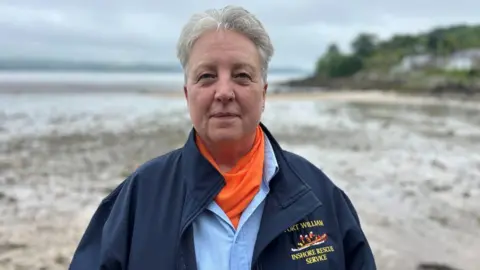 A woman with short grey hair standing on a sandy/muddy beach. She is wearing a blue jacket with a motif which says "inshore rescue service".
