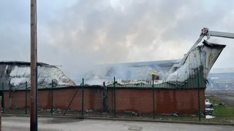 BBC / Andrew Watson A firefighter stands in the collapsed roof of a single storey warehouse building. There is still smoke rising out of the building.