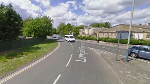 A section of roundabout with a van and two cars in transit and another car leaving. Trees, a grass verge and a number of low buildings are present in the background.